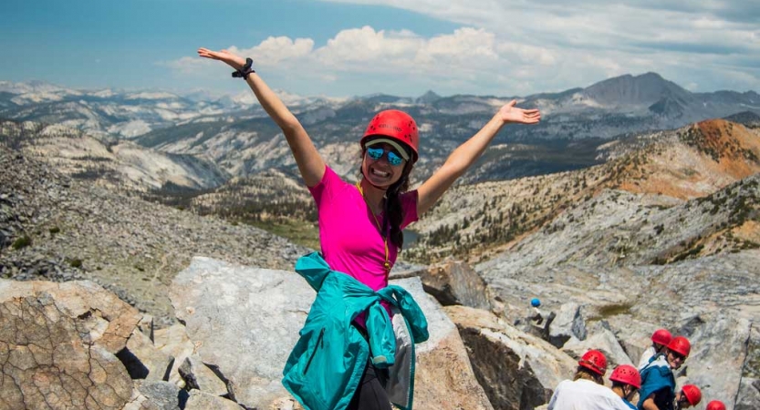 A person wearing a helmet stands and smiles on high ground, with a vast mountain landscape below. 